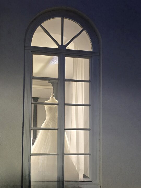 Beleuchtetes Rundbogenfenster der Herzspinnerei bei Nacht, dahinter ein Brautkleid auf einer Büste